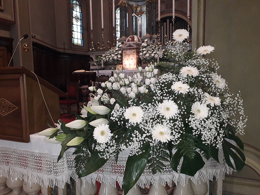 In primo piano una composizione di gerbere bianche velo da sposa, lilium bianchi e ranuncoli bianci con felci mentre dullo sfondo si intravede altare con le stesse composizioni floreali