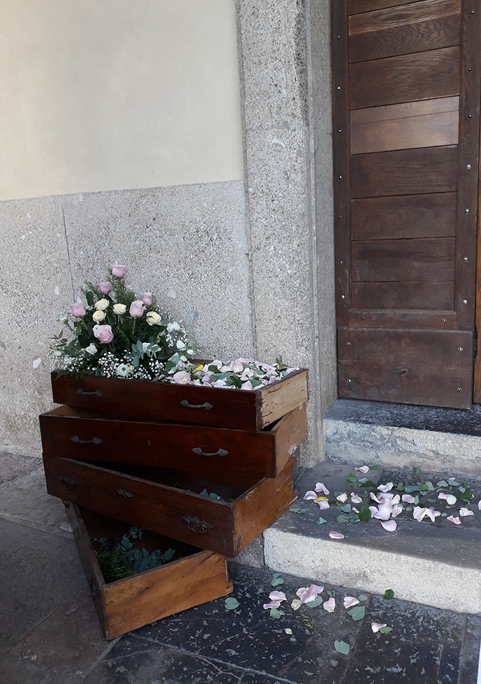 Addobbo esterno ad una chiesetta per la celebrazione di un matrimonio formato da cassetti antichi uno sopra l'altro con all'interno petali colorati rosa bianchi ed una composizione floreale di rose bianche rosa con velo da sposa e foglie verdi