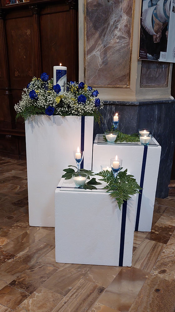 composizione composta da tre basi bianche con nastro blu  una composizione floreale con rose blu velo da sposa e felci  oltre a bicchieri e ciotoline con candele accese bianche e blu